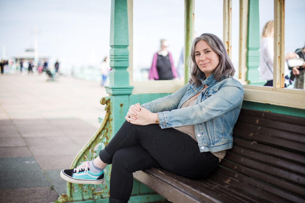 Aimee dressed in denim jacket and black jeans on a bench by the sea front